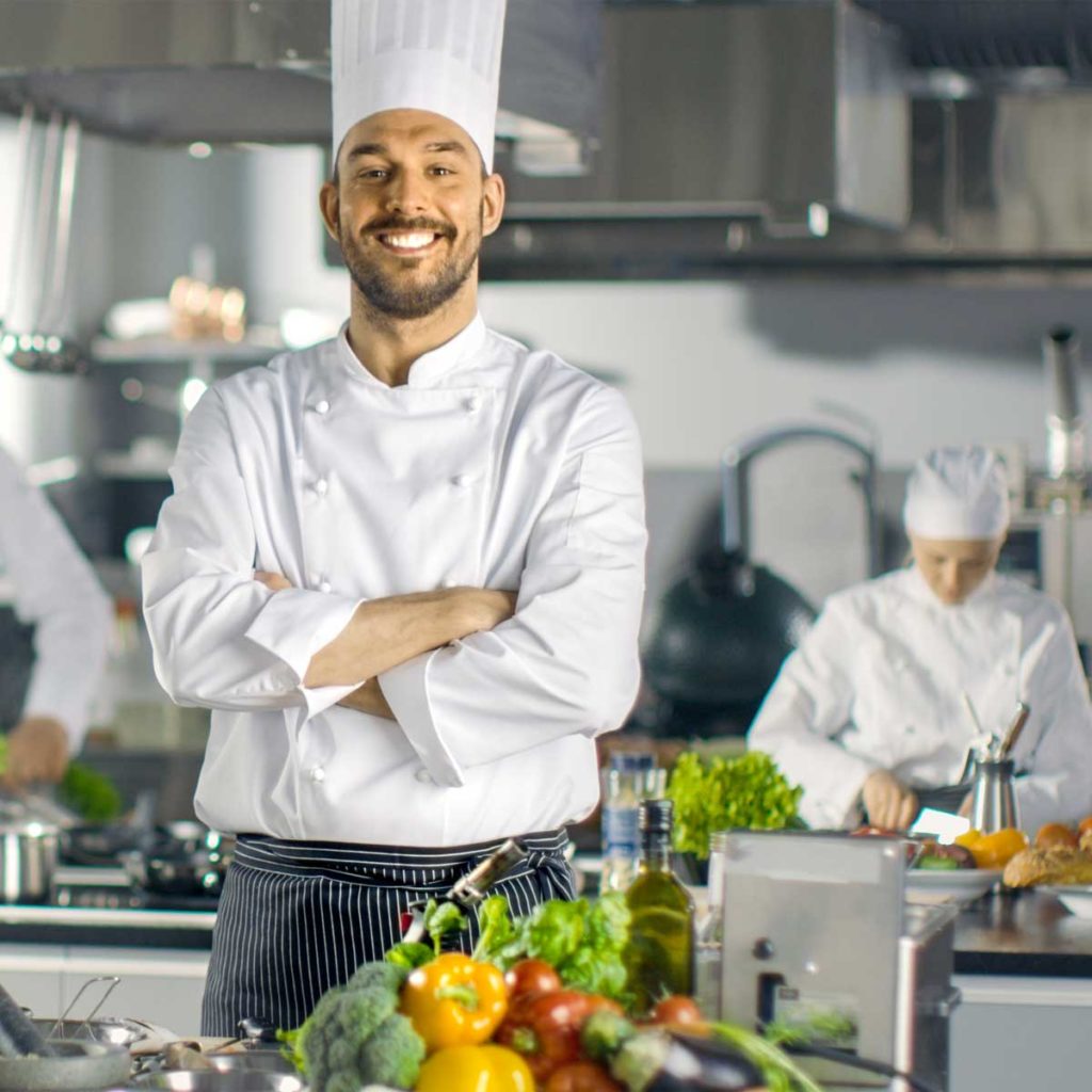 Famous Chef of a Big Restaurant Crosses Arms and Smiles in a Modern Kitchen. His Staff in Working in the Background.